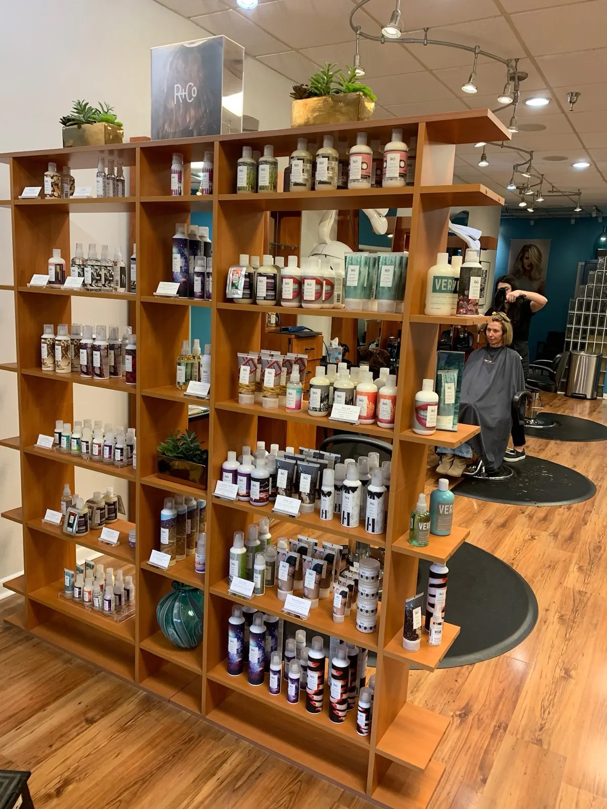 The full R+Co retail shelf inside Ciao Bella Salon, with a guest in the chair behind